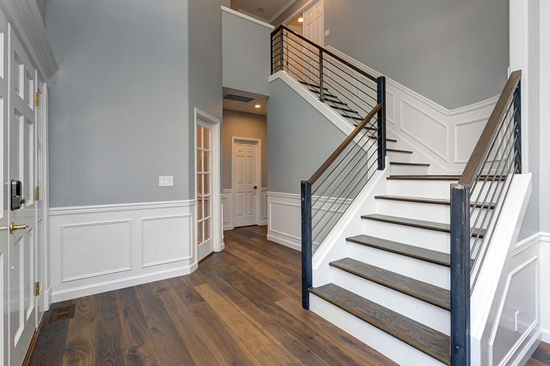 A modern home entryway with light gray walls, white trim, dark wood flooring, and a staircase with dark wood steps and black metal railings, leading to an upper hallway. Multiple white doors are visible.
