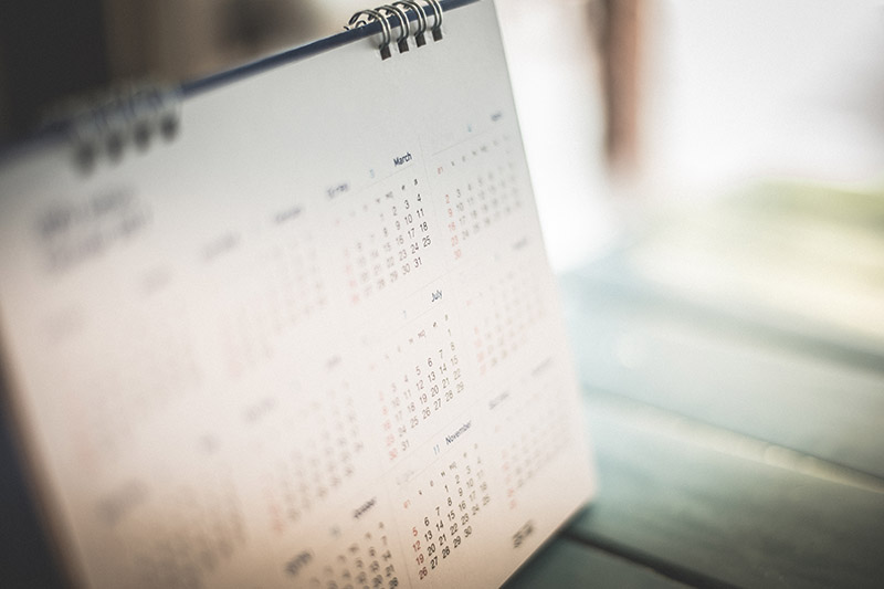 A blurred close-up of a standing desk calendar displaying months and dates, placed on a green wooden surface with soft natural light in the background.