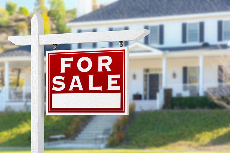 Learning Center 23 A red and white For Sale sign is posted in front of a blurred, large two-story suburban house with white siding, black shutters, and a front porch.