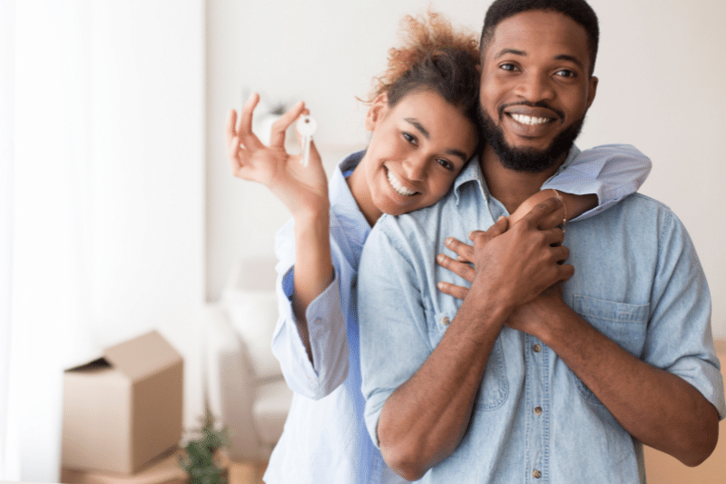 A smiling couple stands close together in a bright room, with the woman holding up a key. Cardboard boxes are in the background, suggesting they have just moved into a new home.
