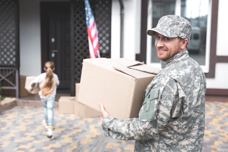 A man in military uniform smiles while holding a cardboard box outside a house. An American flag is near the door, and a young girl carrying a box walks ahead of him toward the house.