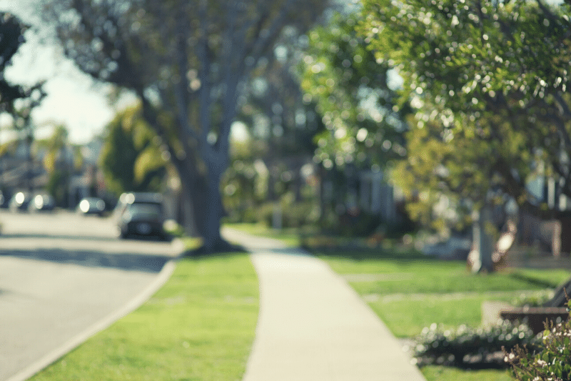 A sunlit suburban sidewalk lined with green grass and trees, with houses and a parked car visible in the background. The image is softly out of focus, creating a peaceful, dreamy atmosphere.