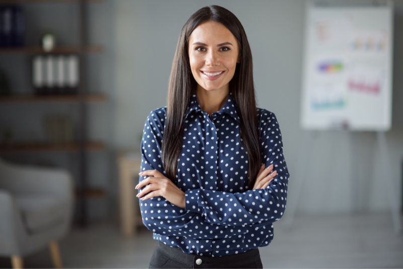 A woman with long dark hair, wearing a navy blue polka dot shirt and black pants, stands confidently with arms crossed and smiles in an office setting. A blurred flipchart and shelves are visible in the background.