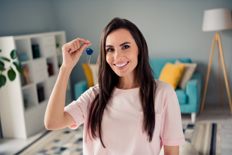 A smiling woman with long dark hair holds up a key indoors, standing in a stylish living room with a blue sofa, yellow pillows, a bookshelf, and a floor lamp in the background.