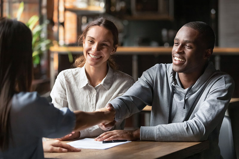 A smiling man and woman sit at a table, shaking hands with a third person whose back is to the camera. Papers and a pen are on the table, suggesting a meeting or agreement in a bright, casual setting.