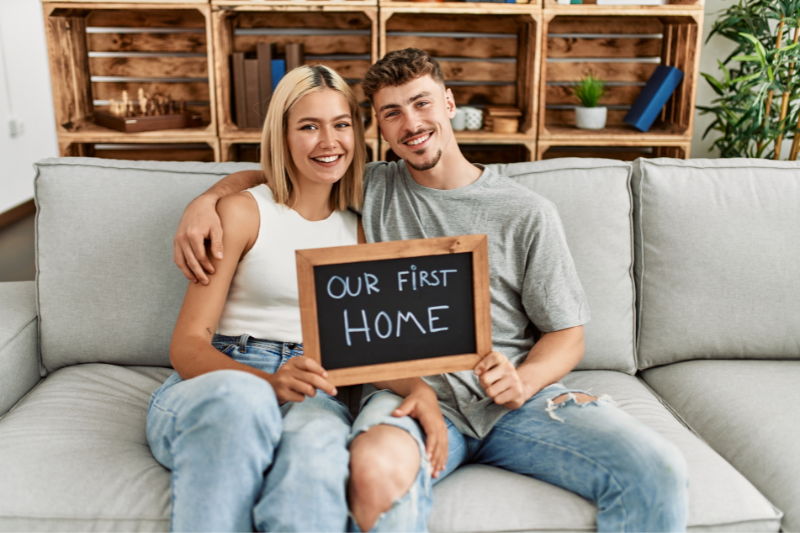 A smiling couple sits on a couch holding a chalkboard sign that reads “Our First Home,” with wooden shelves and plants in the background.