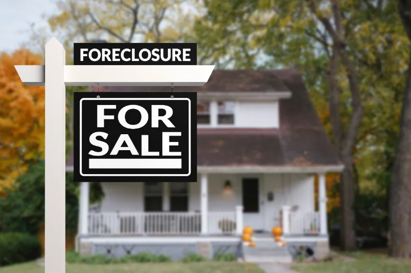 A white post with Foreclosure and For Sale signs stands in front of a white house with a porch, surrounded by trees with autumn leaves. The house appears to be vacant.