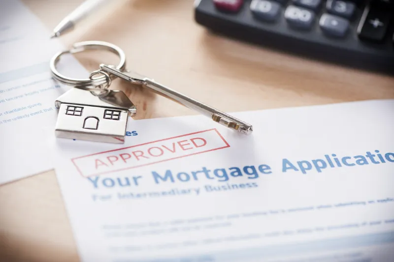 Close-up of a mortgage application form with an APPROVED stamp, a house-shaped keychain, a key, and part of a calculator visible, symbolizing home loan approval and real estate purchase.