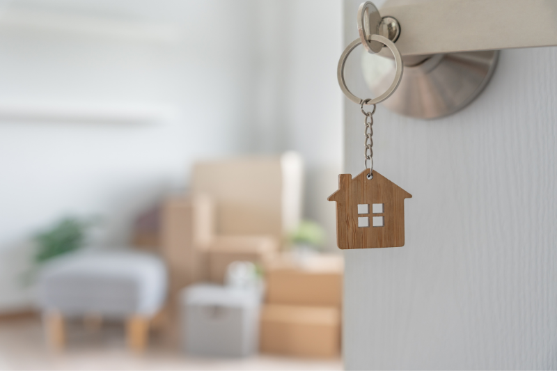 A house-shaped keychain hangs from a key in a door, with packed boxes and furniture visible in a softly focused room, suggesting moving into a new home.