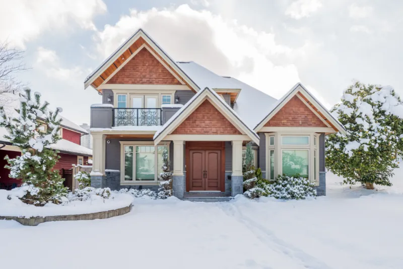 A two-story house with a peaked roof and large front windows is surrounded by snow-covered trees and yard under a bright, partly cloudy sky.