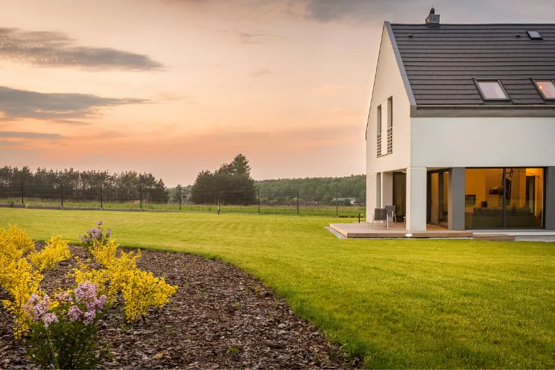 Modern house with large windows and sloped roof, set on a spacious green lawn with yellow and purple flowers, under a cloudy sunset sky, with trees and a fence in the background.