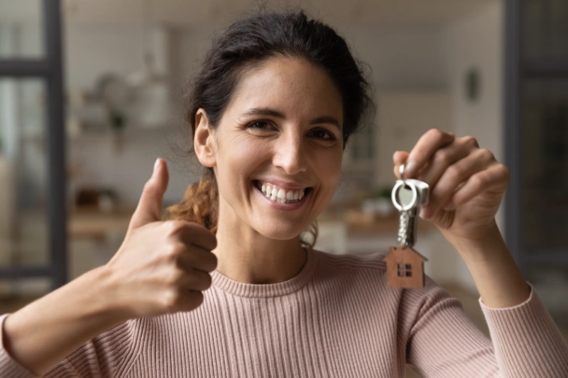Smiling woman holding a house-shaped keychain and giving a thumbs-up, standing indoors in a brightly lit room.